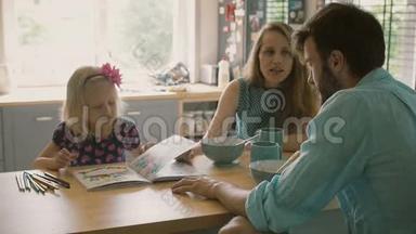 当她的父母在厨房的餐桌上吃早餐时，小女孩正在给她的父母看她的彩色书。 慢节奏